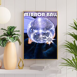 May include: A poster with the text "MIRROR BALL" in white letters on a blue background. A large silver disco ball is in the center of the poster, partially obscured by a person's hand. The text "I'LL SHOW YOU EVERY VERSION OF YOURSELF TONIGHT!" is in a white starburst shape.