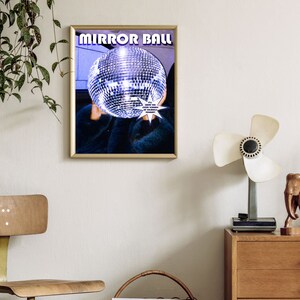 May include: A framed poster with the text "MIRROR BALL" in bold, white letters on a purple background. A silver disco ball is in the center of the poster, partially obscured by a person's hand. The text "I'LL SHOW YOU EVERY VERSION OF YOURSELF" is in white letters on a starburst shape.