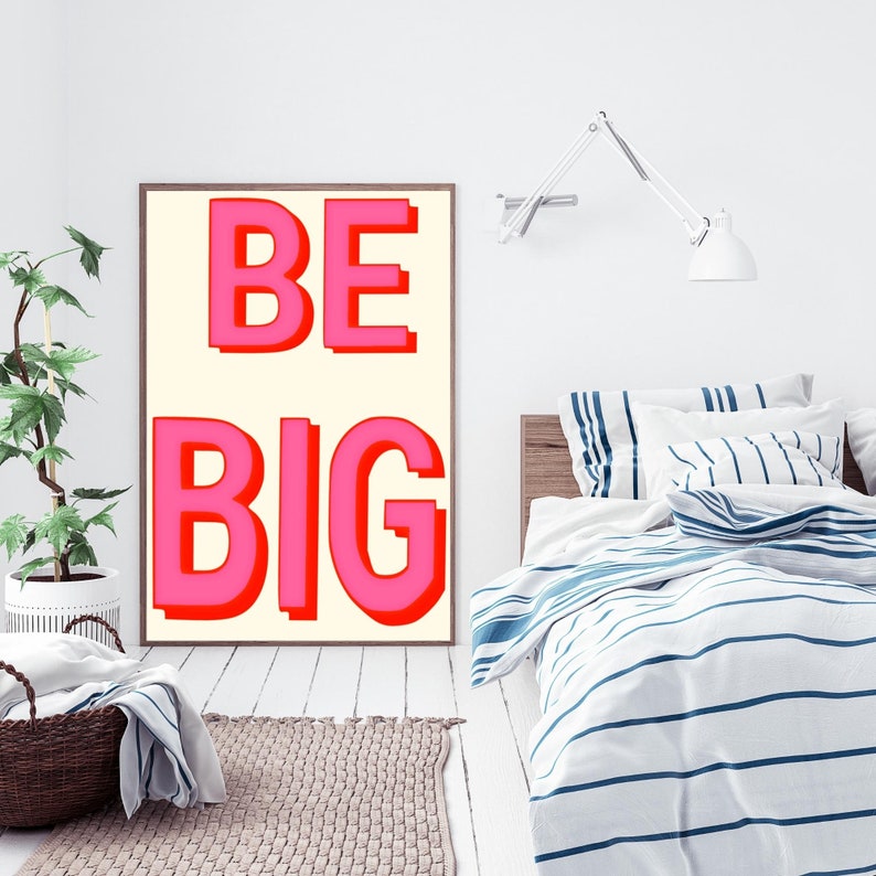 May include: A white framed poster with the words "BE BIG" in pink and red letters on a cream background. The poster is hanging on a white wall above a bed with a white and blue striped duvet cover.