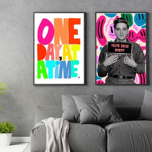 May include: Two framed prints, one with a colorful graphic of the phrase "One Day At A Time" and the other with a black and white photo of Elvis Presley holding a sign that reads "Young Doing Great". The background is a gray wall with a gray couch in the foreground.