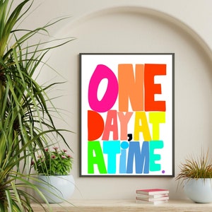 May include: A framed print with the text "ONE DAY AT A TIME." in colorful, bold letters. The letters are in different colors, including pink, orange, red, yellow, green, blue, and purple.