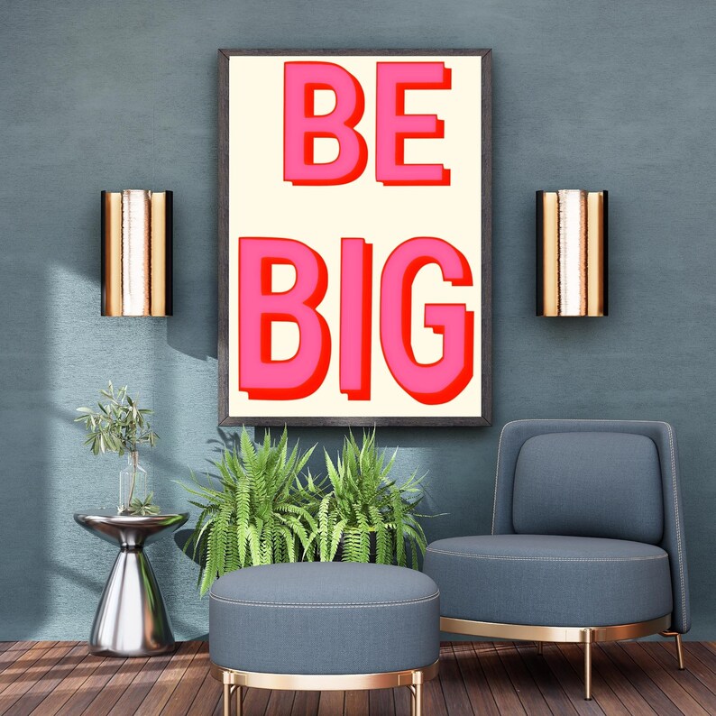 May include: A framed print with the words "BE BIG" in pink letters on a cream background. The print is hanging on a blue wall above a blue armchair and a small round ottoman.