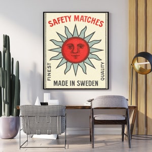 May include: Framed vintage-style poster with the text "SAFETY MATCHES" above a red sun face with blue rays. The poster also includes the text "FINEST QUALITY" and "MADE IN SWEDEN". The poster is displayed on a white wall.