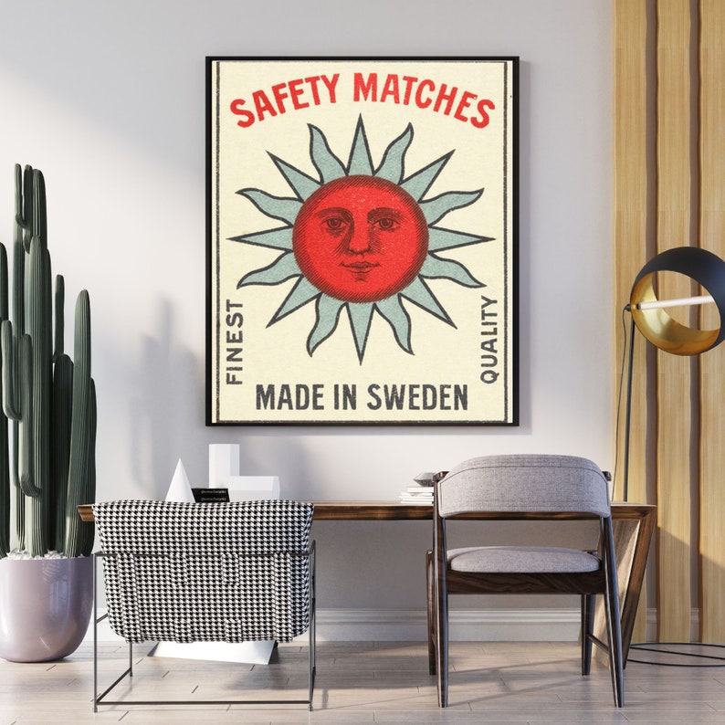 May include: Framed vintage-style poster with the text "SAFETY MATCHES" above a red sun face with blue rays. The poster also includes the text "FINEST QUALITY" and "MADE IN SWEDEN". The poster is displayed on a white wall.