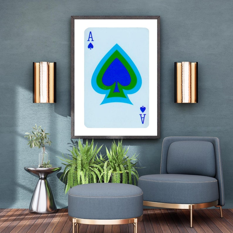 May include: A framed print of an ace of spades playing card with a blue, green, and black spade design. The card is on a light blue background. The print is hanging on a blue wall with two gold wall sconces on either side.