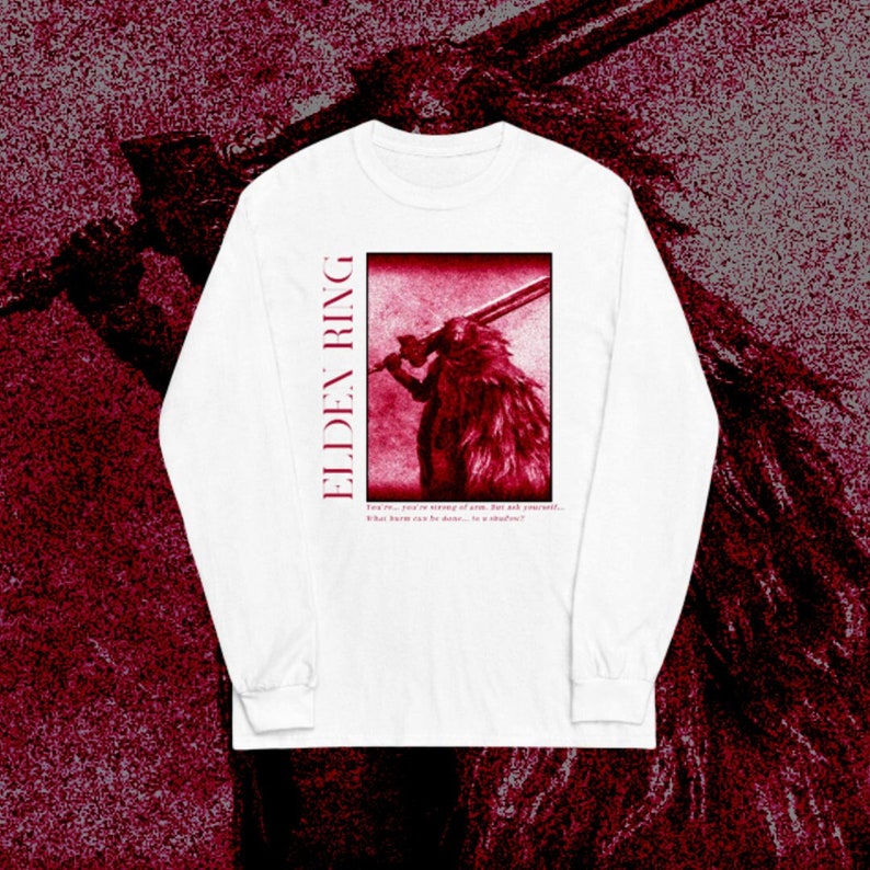 May include: White long-sleeve t-shirt with a red graphic of a character from the video game Elden Ring. The graphic features a character holding a sword and the text "ELDEN RING". The text below the graphic reads "I see me... you are yearning of some... but not yourself... What harm can be done... to a shadow?"