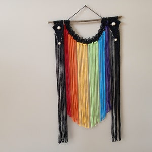 Puede incluir: Un tapiz de macramé arcoíris con hilo negro y una varilla de madera. El tapiz presenta un arcoíris de colores, incluyendo rojo, naranja, amarillo, verde, azul y morado.