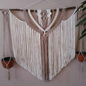 May include: A macrame wall hanging with a geometric design, featuring two hanging plant pots. The wall hanging is made with natural fibres in shades of beige and white.