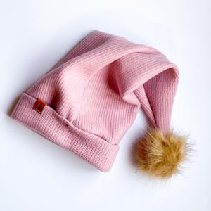 May include: A pink knitted beanie with a brown faux fur pom pom. The beanie has a small brown leather tag with the word "Kids" on it.