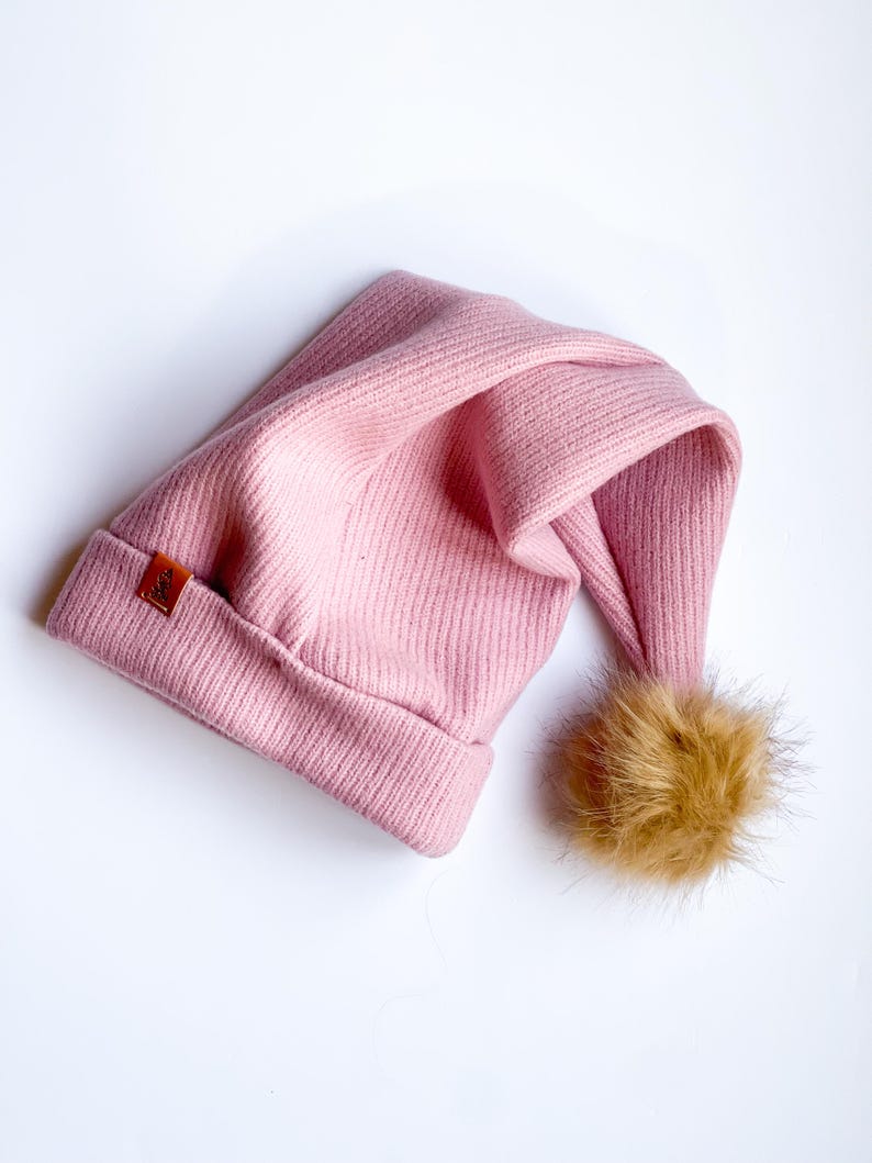 May include: A pink knitted beanie with a brown faux fur pom pom. The beanie has a small brown leather tag with the word "Kids" on it.