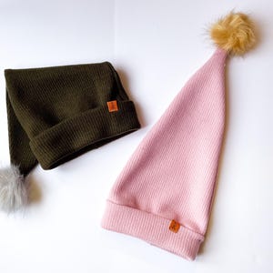May include: Two knitted hats with pom-poms. One hat is olive green with a gray pom-pom and the other is pink with a brown pom-pom.