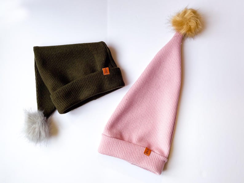 May include: Two knitted hats with pom-poms. One hat is olive green with a gray pom-pom and the other is pink with a brown pom-pom.