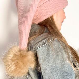 May include: A pink knit beanie with a long, pointed top and a brown faux fur pom pom. The beanie is worn by a person with long, blonde hair.