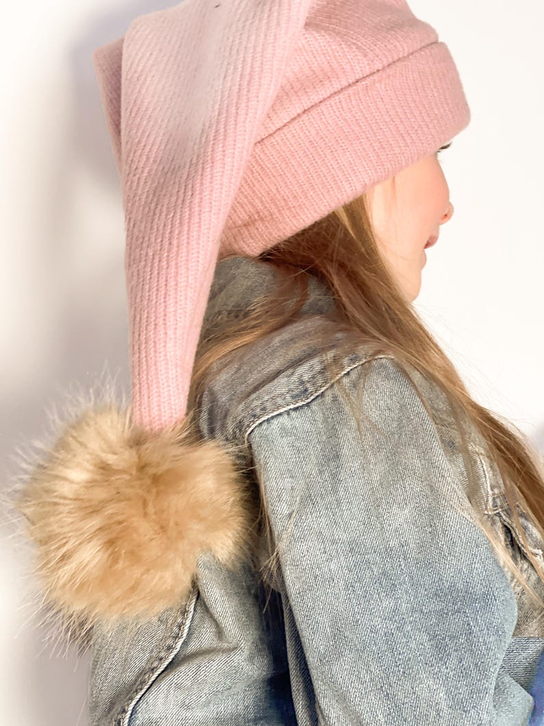 May include: A pink knit beanie with a long, pointed top and a brown faux fur pom pom. The beanie is worn by a person with long, blonde hair.