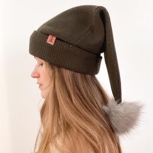 May include: A dark green knit beanie with a long pointed tail and a small faux fur pom-pom. The beanie has a brown leather patch with the word "Cole" on it.