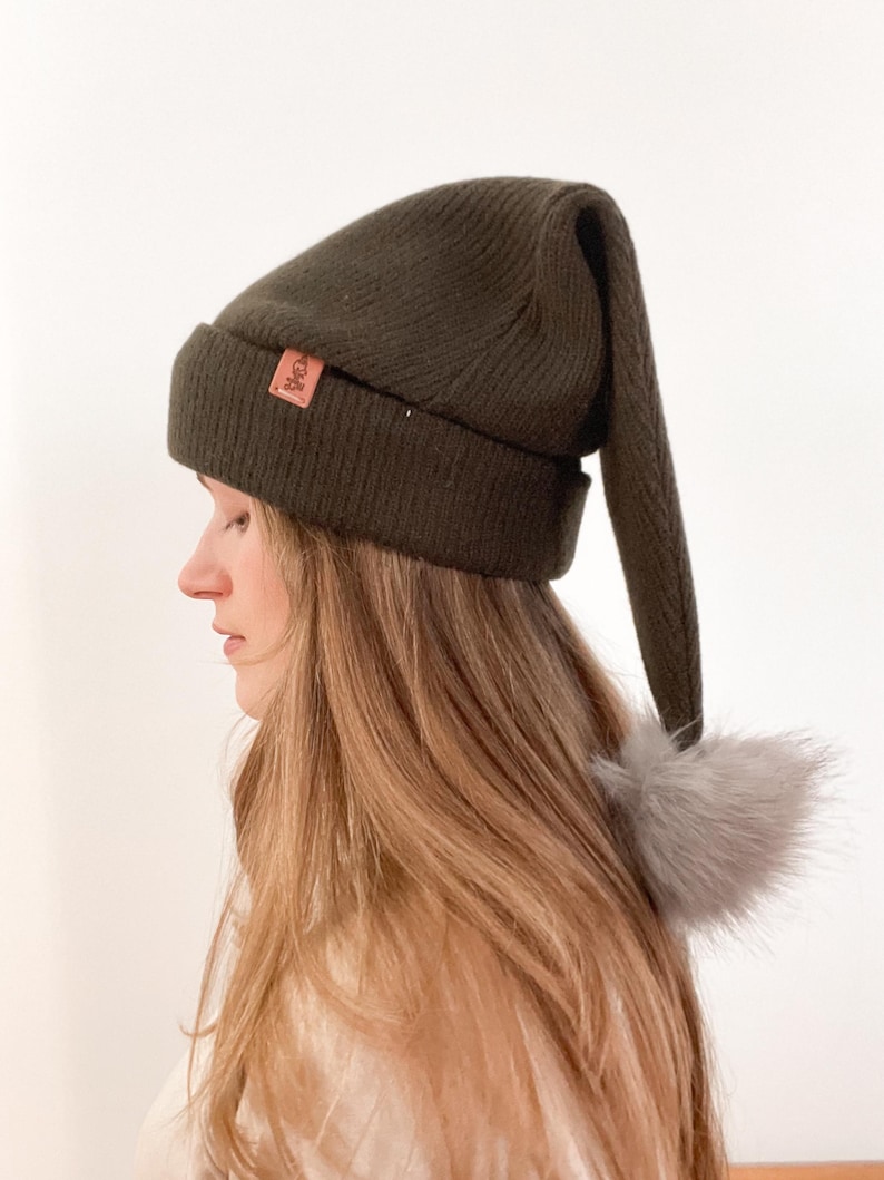 May include: A dark green knit beanie with a long pointed tail and a small faux fur pom-pom. The beanie has a brown leather patch with the word "Cole" on it.