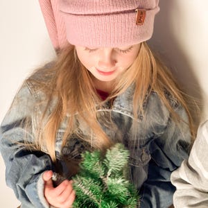 May include: A young girl wearing a pink knit beanie with a brown leather tag is holding a small potted green artificial Christmas tree.