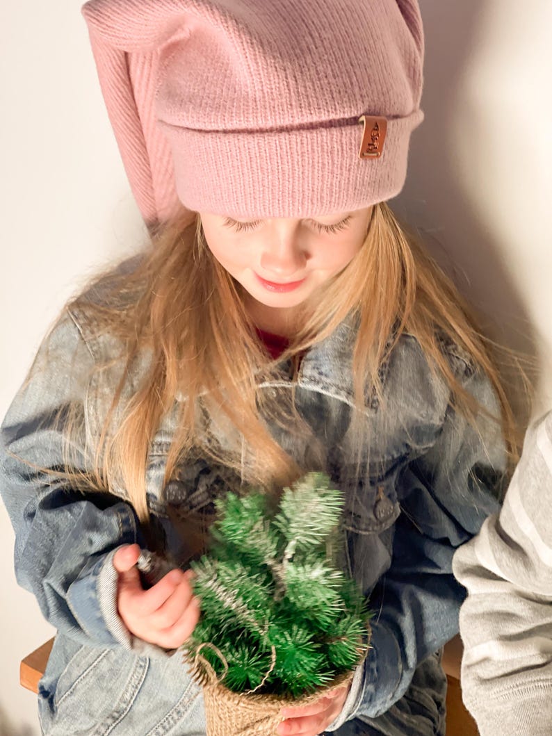 May include: A young girl wearing a pink knit beanie with a brown leather tag is holding a small potted green artificial Christmas tree.