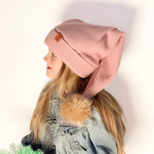 May include: A pink knit beanie with a long tail, featuring a brown leather tag with the word "N.K." on it. The beanie is worn by a person with long blonde hair and a denim jacket.