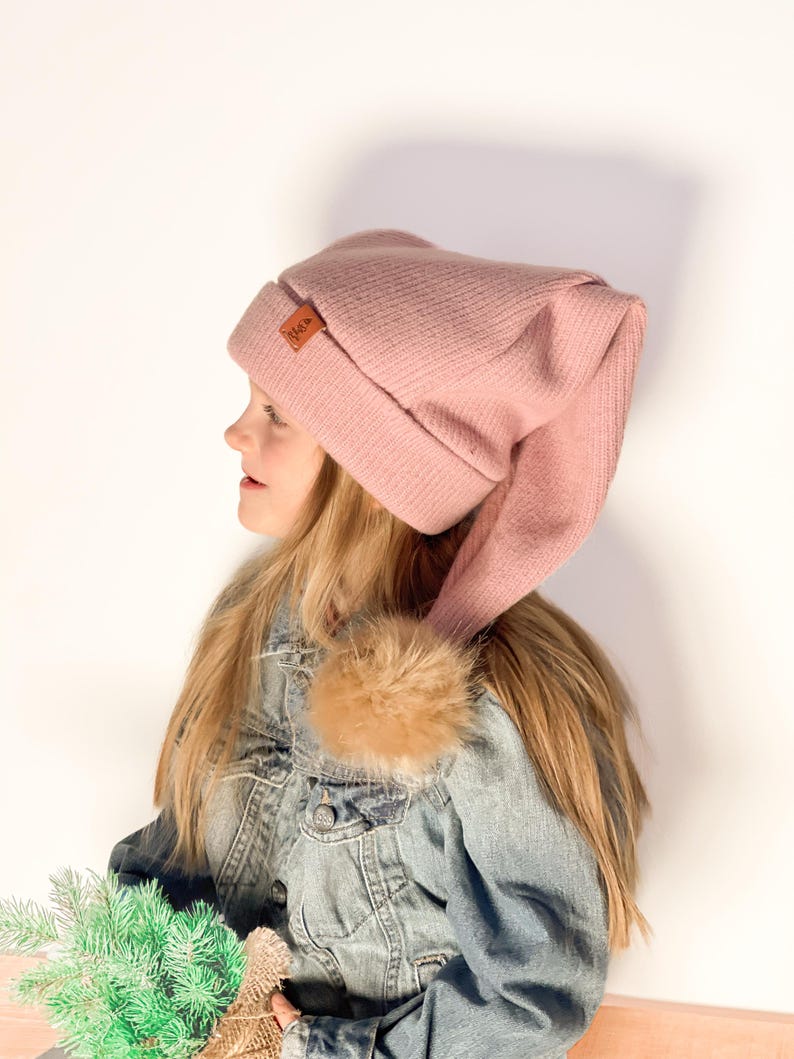 May include: A pink knit beanie with a long tail, featuring a brown leather tag with the word "N.K." on it. The beanie is worn by a person with long blonde hair and a denim jacket.
