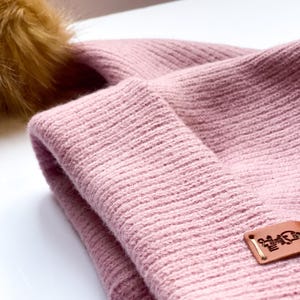 May include: A pink knitted beanie with a brown faux fur pom pom. The beanie has a brown leather patch with the text "Little  Bear" on it.