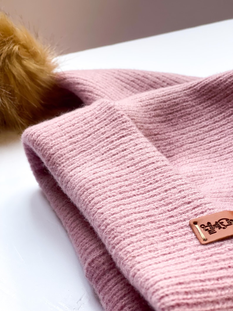 May include: A pink knitted beanie with a brown faux fur pom pom. The beanie has a brown leather patch with the text "Little  Bear" on it.