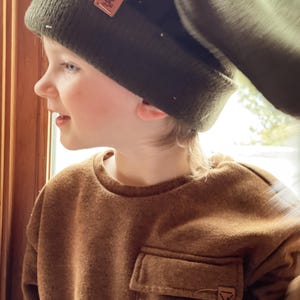 May include: A young person wearing a brown fleece sweatshirt with a patch pocket and a green beanie with a brown leather label. The beanie has a fold-over flap.
