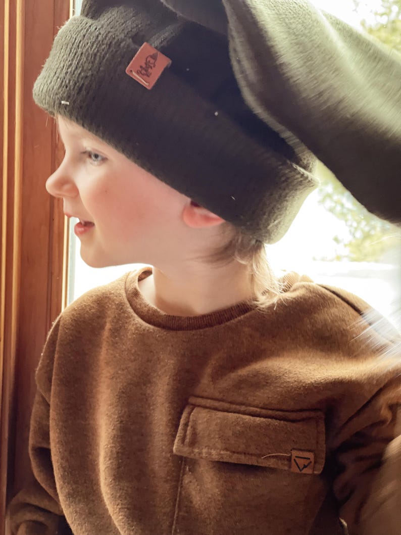 May include: A young person wearing a brown fleece sweatshirt with a patch pocket and a green beanie with a brown leather label. The beanie has a fold-over flap.