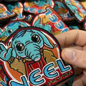 Puede incluir: Un parche bordado azul y naranja con un personaje de dibujos animados y la palabra "NEEL" en letras azules y blancas.