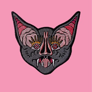 Può includere: Toppa ricamata raffigurante un volto stilizzato di pipistrello. Il pipistrello ha un corpo grigio, dettagli rosa e rossi e uno sfondo rosa. La toppa ha un aspetto dettagliato e strutturato.