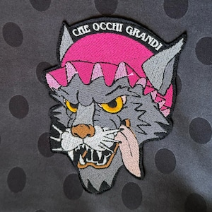 May include: Embroidered patch featuring a stylized wolf head with yellow eyes, a pink hat, and a protruding tongue. The patch has the text "CHE OCCHI GRANDI" in a curved font. The background is gray with black polka dots.