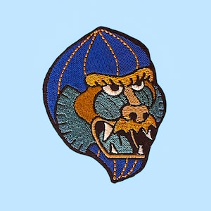 May include: Embroidered patch featuring a gorilla face with a blue, gold, teal, and black color scheme. The gorilla's face is detailed with white and gold thread, and the patch has a black border. The patch is set against a light blue background.