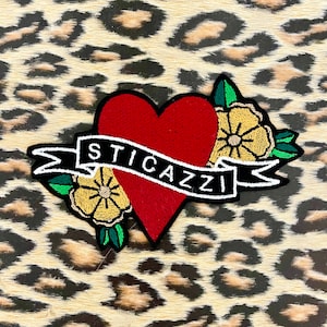 Può includere: Toppa ricamata con un cuore rosso e la scritta "STICAZZI" su uno stendardo. La toppa è decorata con fiori gialli e foglie verdi, su uno sfondo con stampa leopardata.