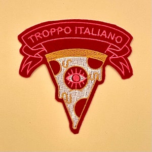 Può includere: Toppa ricamata con un design a fetta di pizza e la scritta "TROPPO ITALIANO" su uno stendardo. La toppa ha uno sfondo rosso con accenti rosa e oro e una crosta di pizza color crema. La fetta di pizza ha un design a occhio.