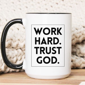 May include: White ceramic mug with a black handle and rim. The mug features the text "WORK HARD. TRUST GOD." in a black, bold font inside a black rectangle. The mug is on a wooden surface.