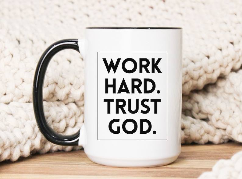 May include: White ceramic mug with a black handle and rim. The mug features the text "WORK HARD. TRUST GOD." in a black, bold font inside a black rectangle. The mug is on a wooden surface.
