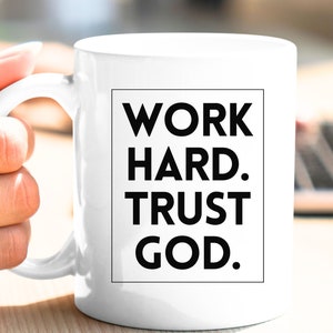 May include: White ceramic mug with the text "WORK HARD. TRUST GOD." in a black, bold font. The mug has a curved handle and is held by a hand. The mug is a standard size for coffee or tea.