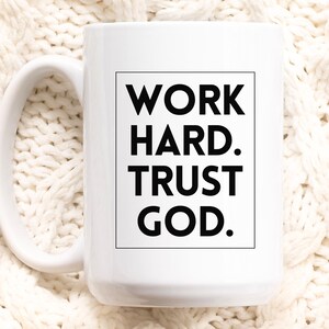 May include: White ceramic mug with a large handle. The mug features the text "WORK HARD. TRUST GOD." in a black, bold font within a black rectangular frame. The mug is set against a white, textured background.