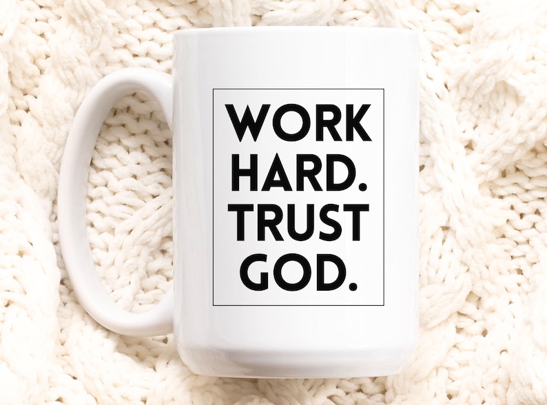 May include: White ceramic mug with a large handle. The mug features the text "WORK HARD. TRUST GOD." in a black, bold font within a black rectangular frame. The mug is set against a white, textured background.