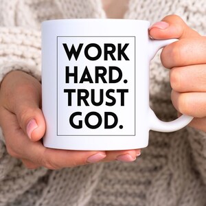 May include: White ceramic mug with the text "WORK HARD. TRUST GOD." in a black, bold font. The mug is held by a person wearing a cream-colored sweater. The mug has a standard handle.