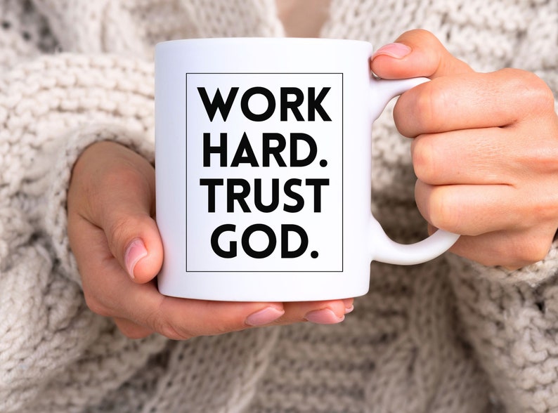 May include: White ceramic mug with the text "WORK HARD. TRUST GOD." in a black, bold font. The mug is held by a person wearing a cream-colored sweater. The mug has a standard handle.