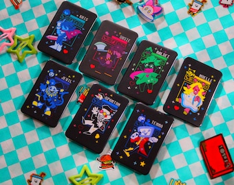 May include: A collection of black rectangular cases, each featuring a different cartoon character design with names like "Kris", "Susie", and "Ralsei". The cases are arranged on a turquoise and white checkered surface, with decorative star-shaped and food-themed accessories.