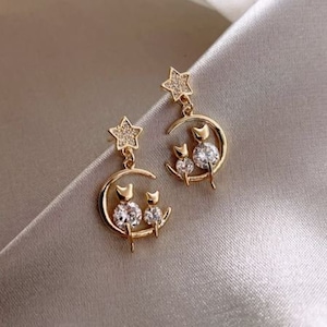 Puede incluir: Pendientes de tono dorado con un diseño de luna creciente y dos gatos sentados en la luna. Los gatos tienen detalles de cristal transparente. Una estrella cuelga de la parte superior de la luna.