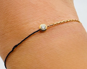 May include: A delicate gold bracelet with a small, round diamond pendant. The bracelet is attached to a black cord on one side and a fine gold chain on the other. The jewelry is worn on a wrist.