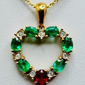 Könnte beinhalten: Eine goldene Halskette mit einem herzförmigen Anhänger. Der Anhänger ist mit ovalen Smaragden, Diamanten und einem zentralen Rubin besetzt. Kette und Anhängerfassung sind aus Gold.