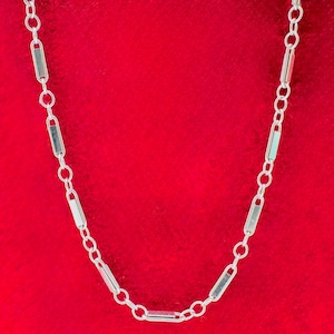 May include: A silver necklace with a delicate chain design. The necklace features a series of rectangular links connected by small circular links. The jewelry is displayed against a vibrant red background, highlighting its metallic shine.