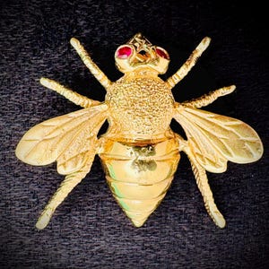 Puede incluir: Broche dorado con forma de abeja, con ojos de piedras preciosas rojas y una corona. El broche tiene alas y patas detalladas, y un cuerpo texturizado. La joya está sobre un fondo oscuro.