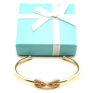 Könnte beinhalten: Ein goldfarbenes Unendlichkeits-Armband mit klaren Zirkonia-Steinen. Das Armband befindet sich in einer hellblauen Geschenkbox mit einem weißen Band.