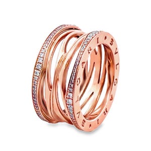 Puede incluir: Anillo de oro rosa con un diseño único y en capas. El anillo presenta múltiples bandas que se entrelazan, creando una apariencia abierta y texturizada. Las bandas exteriores están adornadas con pequeñas piedras preciosas brillantes. El anillo tiene un acabado pulido y una estética lujosa.