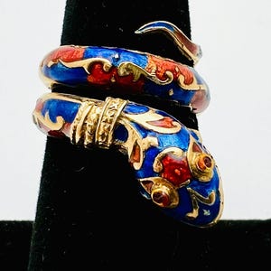 May include: A gold-toned snake ring with blue, red, and gold enamel detailing. The ring is coiled, with the snake's head and tail overlapping. The design features intricate patterns and a polished finish, showcasing a unique jewelry piece.